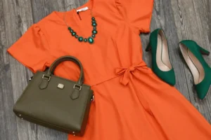 Flatlay of an orange dress paired with green heels, a green handbag, and a green necklace. The vibrant green accessories create a stunning contrast with the orange dress, offering a stylish colour combination for orange dress. Visit yesvsno.net for more outfit inspiration