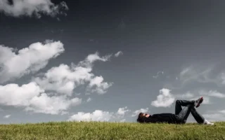 Person lying on the grass under a cloudy sky, reflecting on a peaceful Sunday routine and enjoying nature – YesvsNo.net.