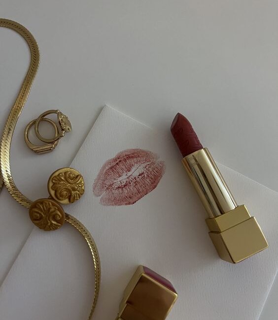 A stylish flat lay featuring a lipstick with a kiss mark on paper, complemented by rings and a necklace. The rich, deep shade of the lipstick represents a chic and timeless makeup look. Trending makeup looks featured on yesvsno.net.