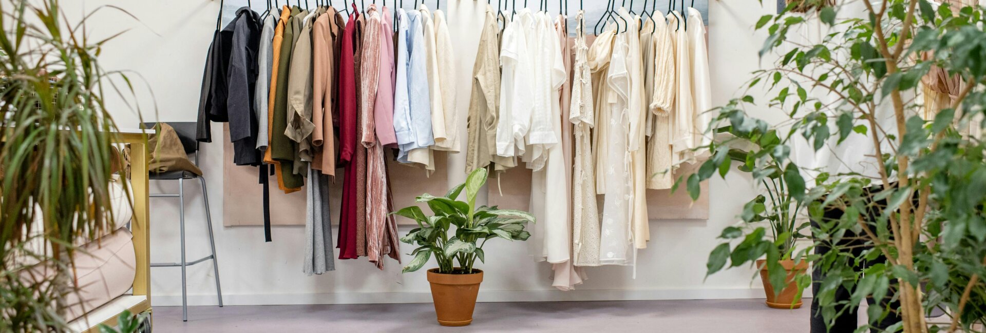 A clean and minimalist boutique space featuring clothing racks with a mix of neutral and earthy-toned garments neatly hung. The scene includes potted green plants placed throughout the room, creating an eco-friendly and serene atmosphere.