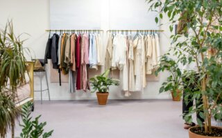 A clean and minimalist boutique space featuring clothing racks with a mix of neutral and earthy-toned garments neatly hung. The scene includes potted green plants placed throughout the room, creating an eco-friendly and serene atmosphere.