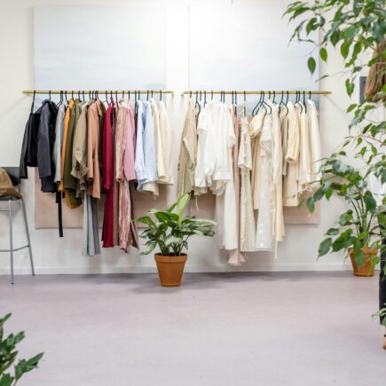 A clean and minimalist boutique space featuring clothing racks with a mix of neutral and earthy-toned garments neatly hung. The scene includes potted green plants placed throughout the room, creating an eco-friendly and serene atmosphere.