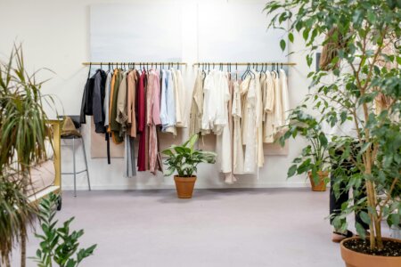 A clean and minimalist boutique space featuring clothing racks with a mix of neutral and earthy-toned garments neatly hung. The scene includes potted green plants placed throughout the room, creating an eco-friendly and serene atmosphere.