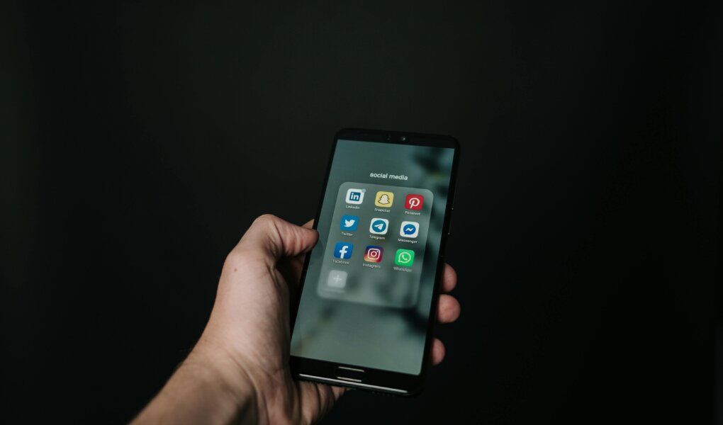 This image shows a hand holding a smartphone against a dark background, with the screen displaying a folder labeled "social media" containing popular app icons such as LinkedIn, Snapchat, Pinterest, Twitter, Telegram, Messenger, Facebook, Instagram, and WhatsApp. The minimalistic composition emphasizes the smartphone and its content, suggesting a focus on social media usage.