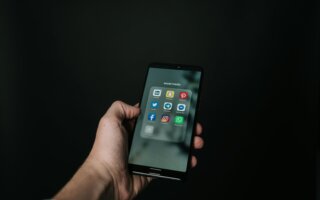 This image shows a hand holding a smartphone against a dark background, with the screen displaying a folder labeled "social media" containing popular app icons such as LinkedIn, Snapchat, Pinterest, Twitter, Telegram, Messenger, Facebook, Instagram, and WhatsApp. The minimalistic composition emphasizes the smartphone and its content, suggesting a focus on social media usage and prompting the question, should you quit social media?