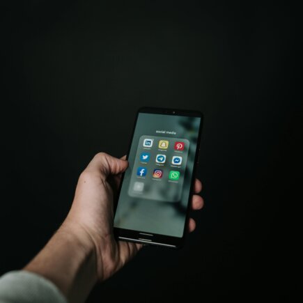 This image shows a hand holding a smartphone against a dark background, with the screen displaying a folder labeled "social media" containing popular app icons such as LinkedIn, Snapchat, Pinterest, Twitter, Telegram, Messenger, Facebook, Instagram, and WhatsApp. The minimalistic composition emphasizes the smartphone and its content, suggesting a focus on social media usage.