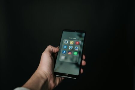 This image shows a hand holding a smartphone against a dark background, with the screen displaying a folder labeled "social media" containing popular app icons such as LinkedIn, Snapchat, Pinterest, Twitter, Telegram, Messenger, Facebook, Instagram, and WhatsApp. The minimalistic composition emphasizes the smartphone and its content, suggesting a focus on social media usage.