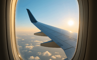 Realistic-view-from-an-airplane-window-during-early-morning-featuring-a-clear-bright-blue-sky-with-scattered-fluffy-white-clouds.-The-airplane-wing-extends-diagonally-across-the-frame-bathed-in-soft-sunlight-casting-a-warm-glow.-The-window-has-a-subtle-reflection-and-the-horizon-is-peaceful-and-clear-with-no-visible-landmarks-emphasizing-the-tranquility-of-a-calm-morning-sky