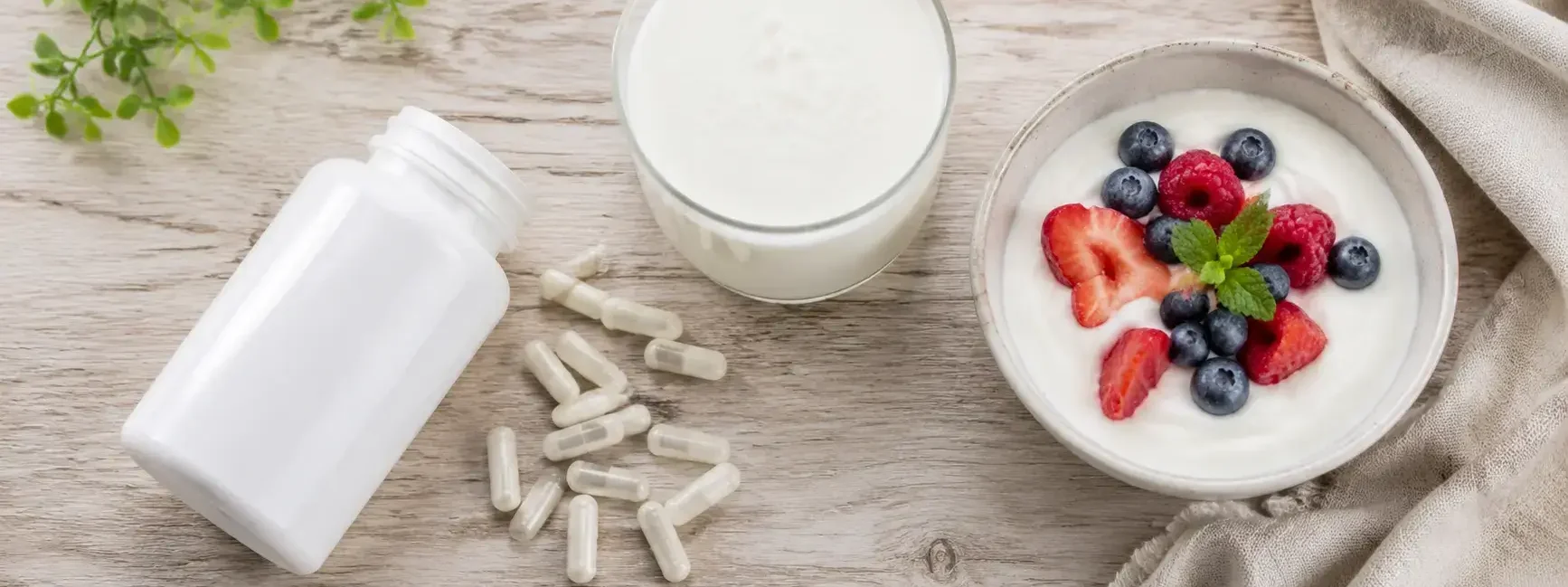 Flat lay of probiotic supplement bottle with capsules spilled beside kefir, yogurt, and berries on a light wooden surface under soft natural lighting — probiotic supplements and fermented foods - yes or no to probiotics every day.