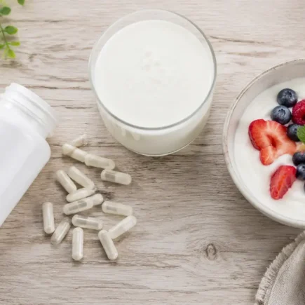Flat lay of probiotic supplement bottle with capsules spilled beside kefir, yogurt, and berries on a light wooden surface under soft natural lighting — probiotic supplements and fermented foods - yes or no to probiotics every day.