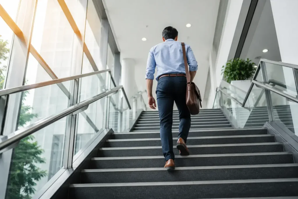 A lifestyle shot of a person taking the stairs in an office building, seen from a low angle looking up, natural building light, business casual outfit, energetic and purposeful feel.