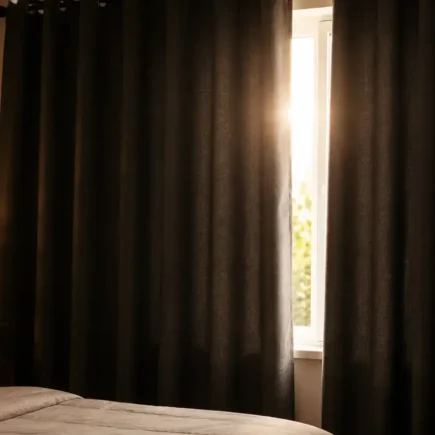 A flat lay of thick dark fabric curtains partially drawn across a bedroom window blocking bright sunlight, soft warm interior lighting, minimal and editorial style. No text overlays.yes or no to blackout curtains for sleep.