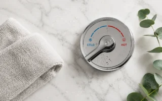 A flat lay of a cold shower temperature dial turned to cold, a small towel, and a sprig of eucalyptus on a clean white marble surface. Crisp, minimal, fresh feel. No text overlays.yes or no to cold showers.
