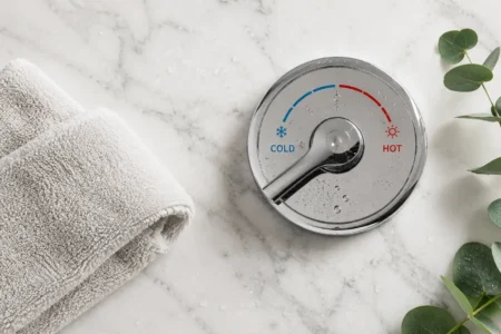A flat lay of a cold shower temperature dial turned to cold, a small towel, and a sprig of eucalyptus on a clean white marble surface. Crisp, minimal, fresh feel. No text overlays.yes or no to cold showers.