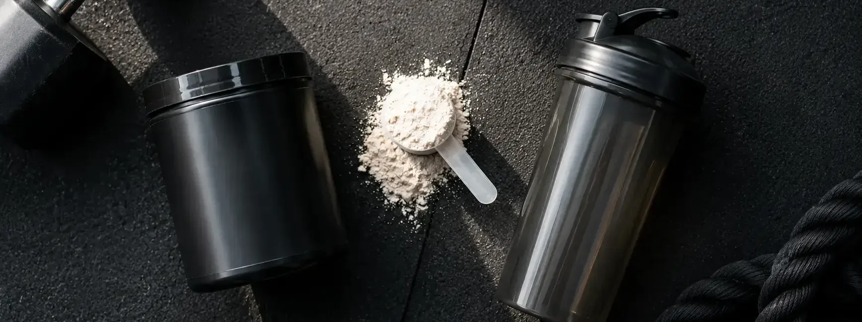 Top-down flat lay of a pre-workout supplement tub, a scoop of powder, and a shaker bottle on a dark gym floor under natural light — pre-workout supplement powder and shaker bottle - yes or no to pre-workout for fat loss.