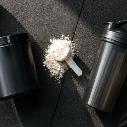 Top-down flat lay of a pre-workout supplement tub, a scoop of powder, and a shaker bottle on a dark gym floor under natural light — pre-workout supplement powder and shaker bottle - yes or no to pre-workout for fat loss.