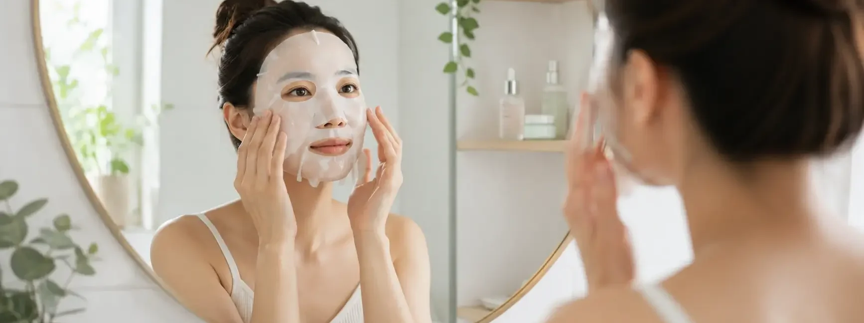A person applying a hydrating sheet mask in a bright, clean bathroom mirror, soft natural light, minimal skincare shelf visible in the background.