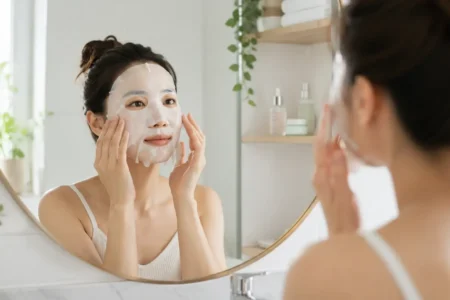 A person applying a hydrating sheet mask in a bright, clean bathroom mirror, soft natural light, minimal skincare shelf visible in the background.