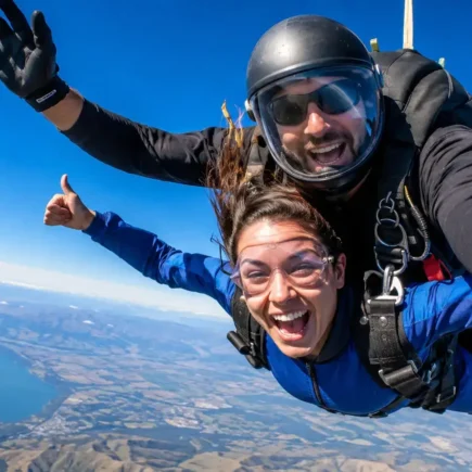 A tandem skydiving pair in freefall against a clear blue sky, visible parachute pack, wide open landscape below, exhilarated expression - yes or no to skydiving featured image.