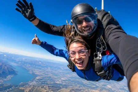 A tandem skydiving pair in freefall against a clear blue sky, visible parachute pack, wide open landscape below, exhilarated expression - yes or no to skydiving featured image.
