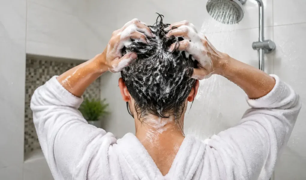 A person washing their hair in the shower, close-up on hands massaging shampoo into the scalp, bright clean bathroom, water droplets visible.yes or no to washing your hair every day featured image.