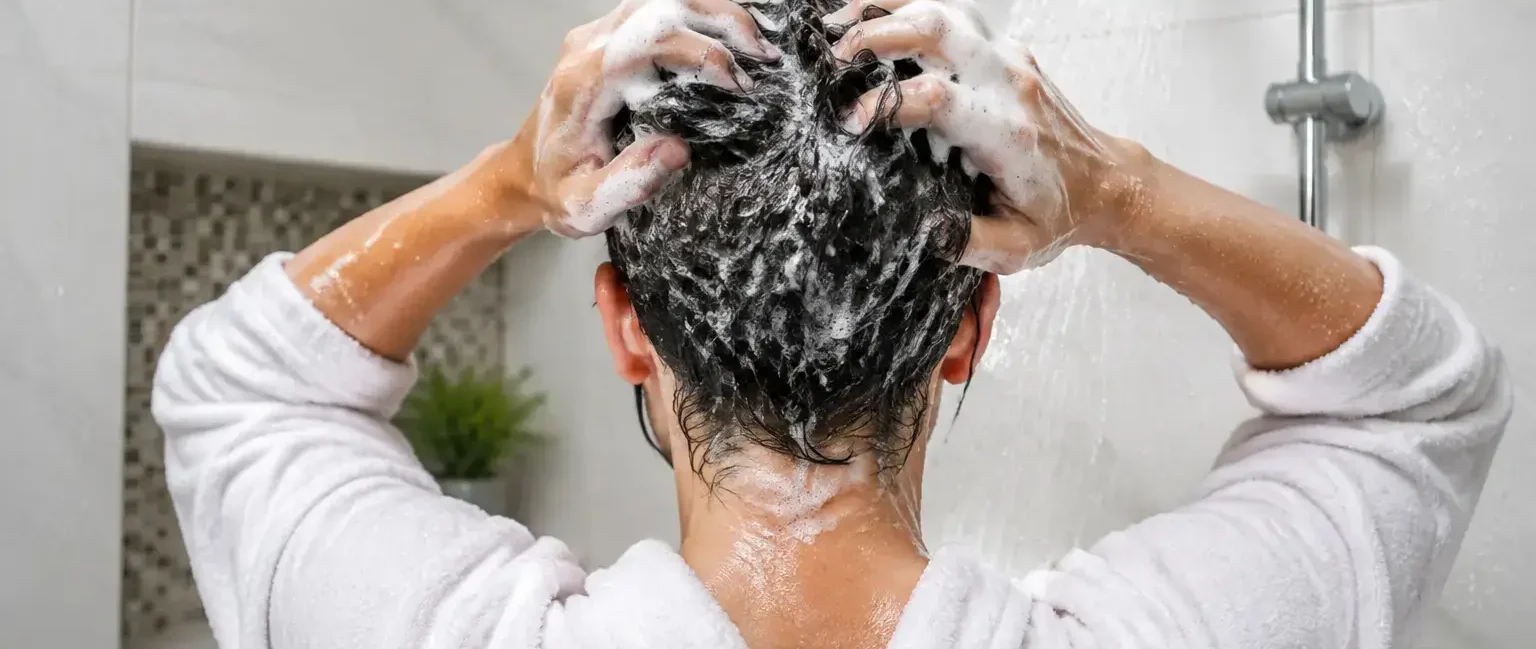A person washing their hair in the shower, close-up on hands massaging shampoo into the scalp, bright clean bathroom, water droplets visible.yes or no to washing your hair every day featured image.