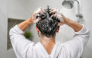A person washing their hair in the shower, close-up on hands massaging shampoo into the scalp, bright clean bathroom, water droplets visible.yes or no to washing your hair every day featured image.