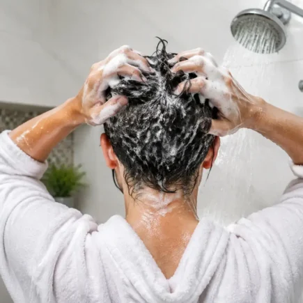 A person washing their hair in the shower, close-up on hands massaging shampoo into the scalp, bright clean bathroom, water droplets visible.yes or no to washing your hair every day featured image.