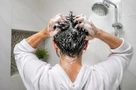 A person washing their hair in the shower, close-up on hands massaging shampoo into the scalp, bright clean bathroom, water droplets visible.yes or no to washing your hair every day featured image.